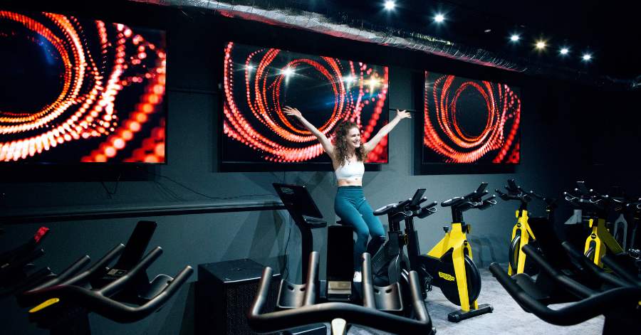 A dynamic spin class image showing a smiling member with arms raised in victory on a stationary bike, set against three large, vibrant digital screens. This captures the high-energy atmosphere at RINTO Fitness, the best gym in Richmond.