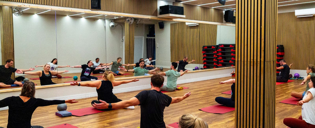 Group fitness class at Rinto Fitness with participants sitting on yoga mats with arms outstretched.